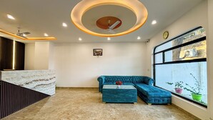 Un hall d’hôtel moderne avec un sol en marbre, un canapé bleu, un ventilateur de plafond et une grande fenêtre ornée de plantes.