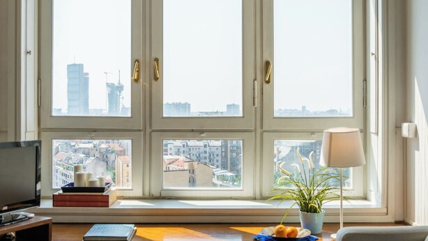 Una habitación con un ventanal grande, televisión, lámpara y una mesita con libros y una planta.