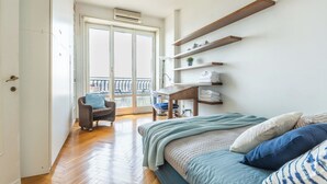 Un dormitorio con cama, escritorio y silla. Cuenta con un balcón y barandilla.