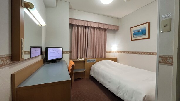 Una habitación de hotel con cama, escritorio, computadora, teléfono y ventana con cortinas.