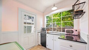 Una cocina con armarios blancos, lavavajillas, microondas y ventana con vista a follaje.