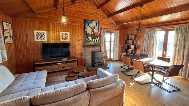 Ein gemütliches Blockhaus-Interieur mit Holzbalkendecke, Kamin, einer Couch, einem Esstisch und einem Fernseher.
