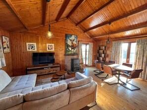 Interior acogedor de una cabaña con techo de madera, chimenea, sofá, mesa de comedor y televisor.
