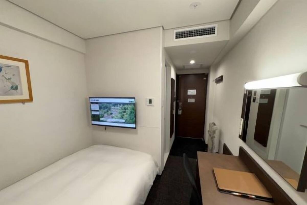 A hotel room with a bed, a desk with a laptop, a TV mounted on the wall, and a door leading to another room.