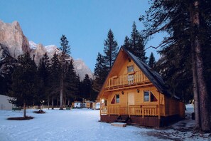 Una cabaña de madera con balcón, rodeada de nieve y árboles, con montañas al fondo.
