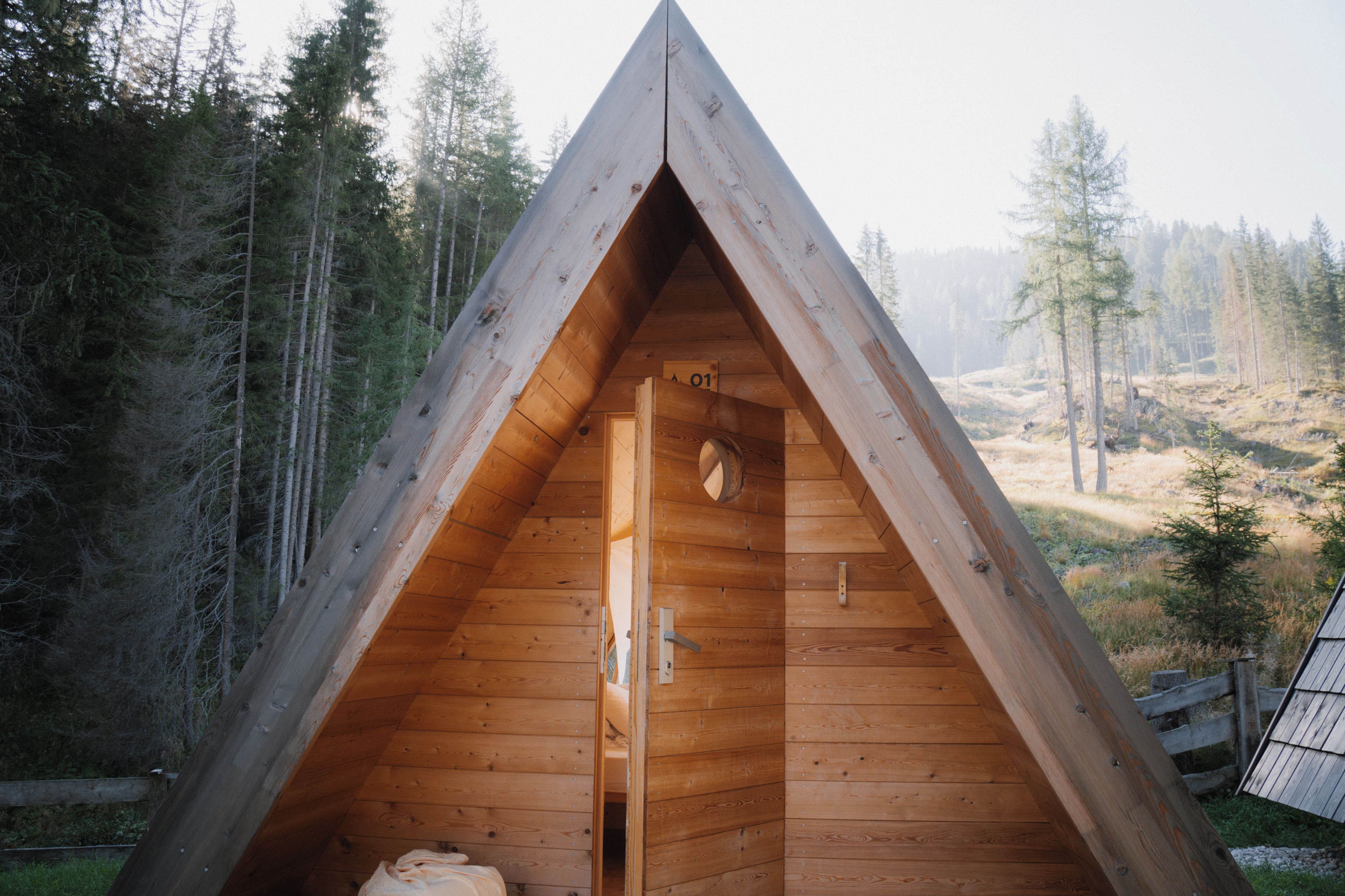 Una cabaña de madera triangular con una puerta y una ventana, rodeada de un bosque.