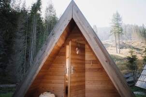 Una cabaña de madera triangular con una puerta y una ventana, rodeada de un bosque.