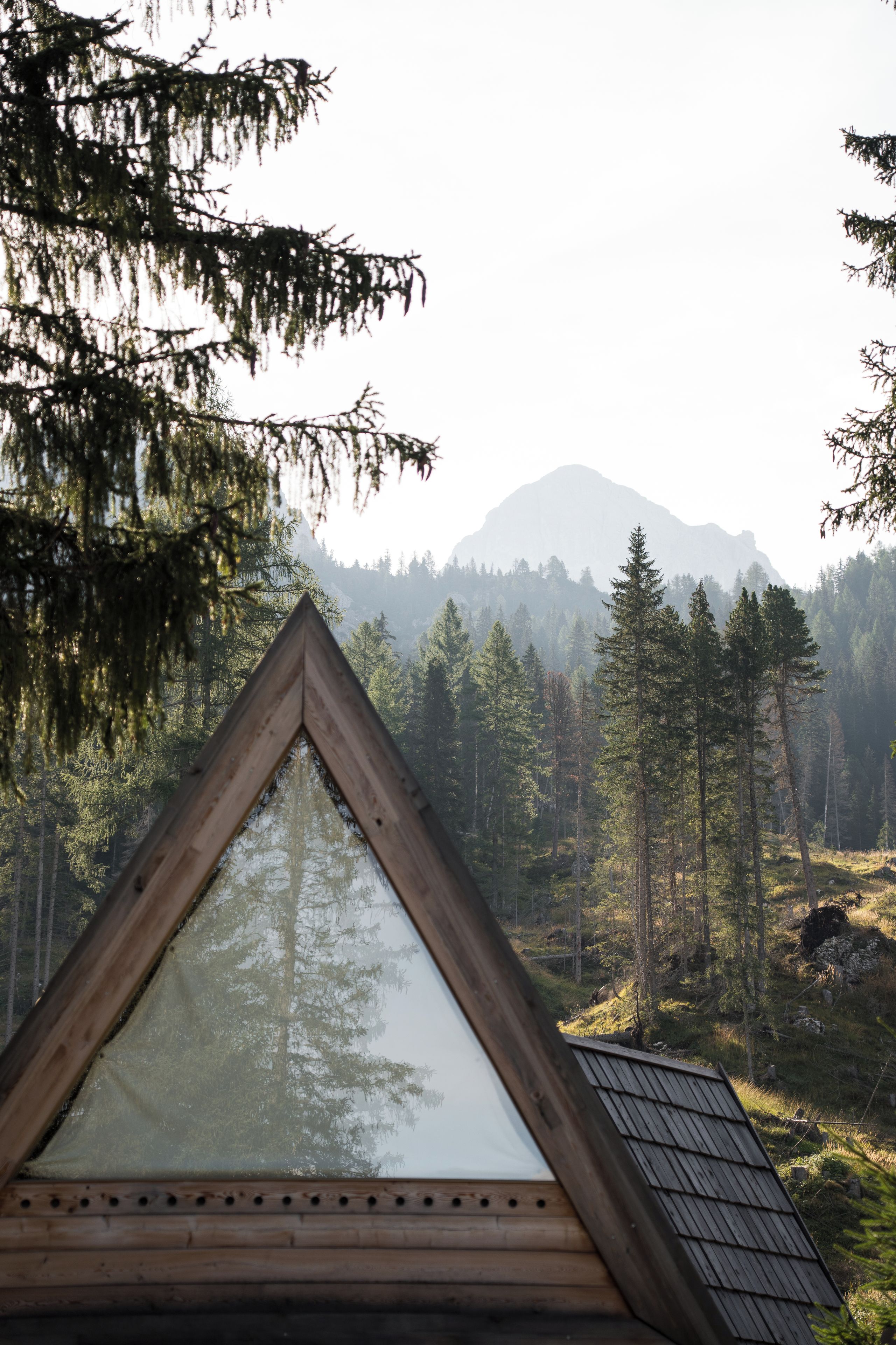 Una cabaña con un ventanal triangular, rodeada de un bosque y una montaña al fondo.
