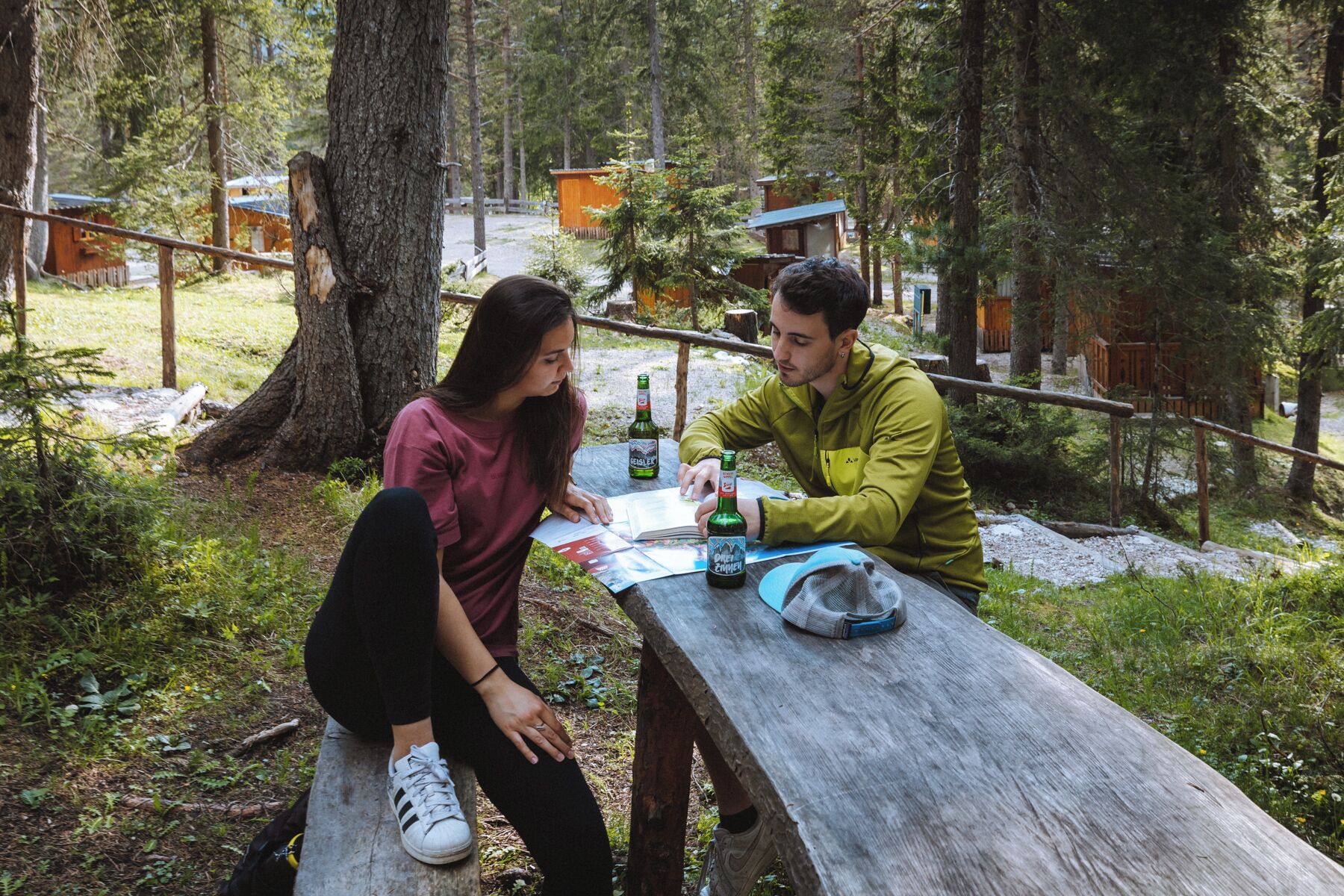 Two people sitting at a wooden picnic table outdoors with trees and cabins in the background.