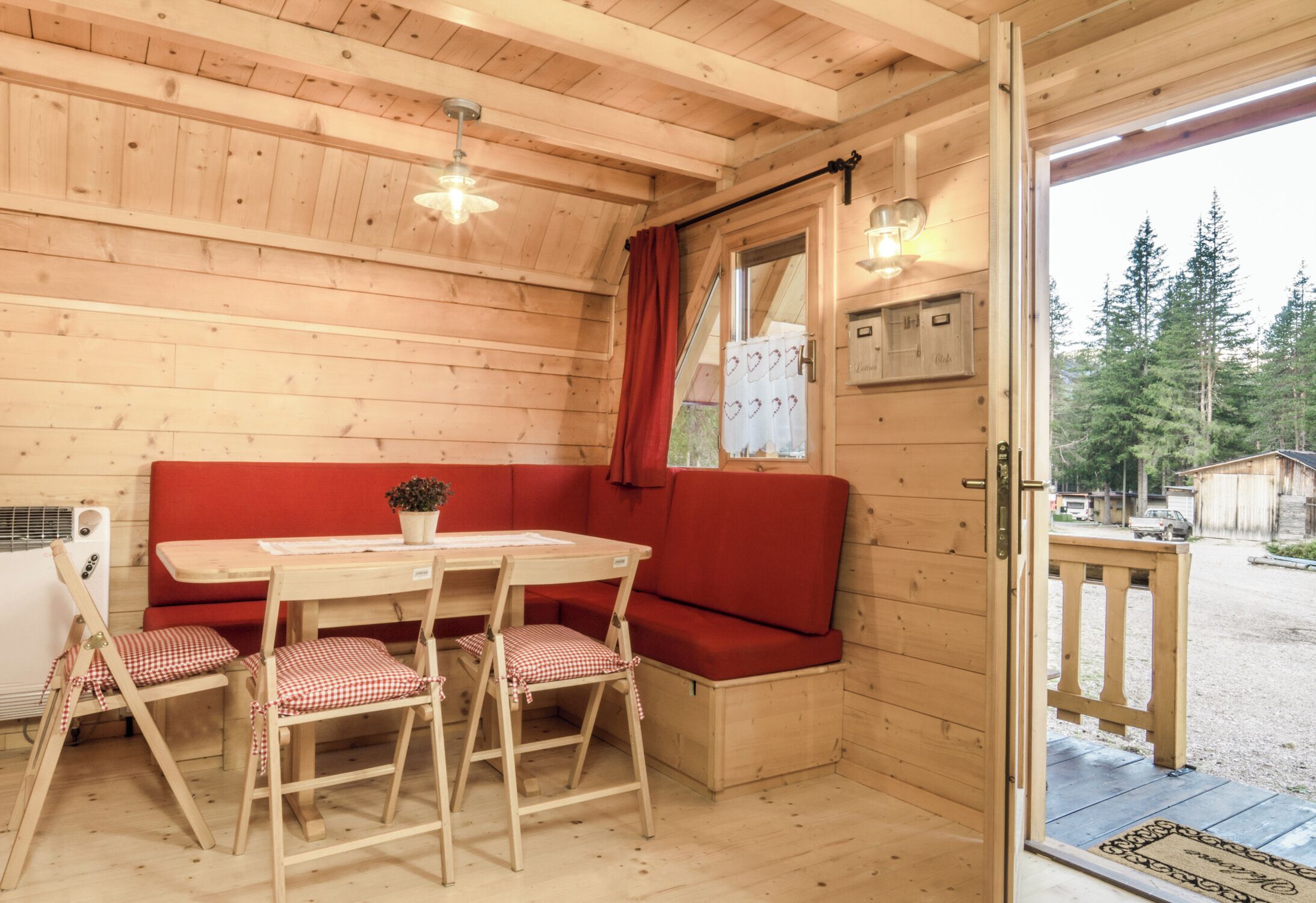 A wooden cabin interior with a dining table, red cushioned bench, and a view of the forest.