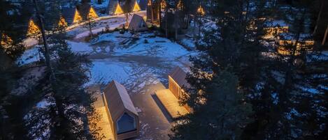 Un bosque cubierto de nieve con varias cabañas de techo triangular iluminadas por luces cálidas.