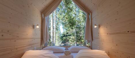 A wooden cabin room with a bed, two pillows, a rolled towel, and a window with a view of trees.