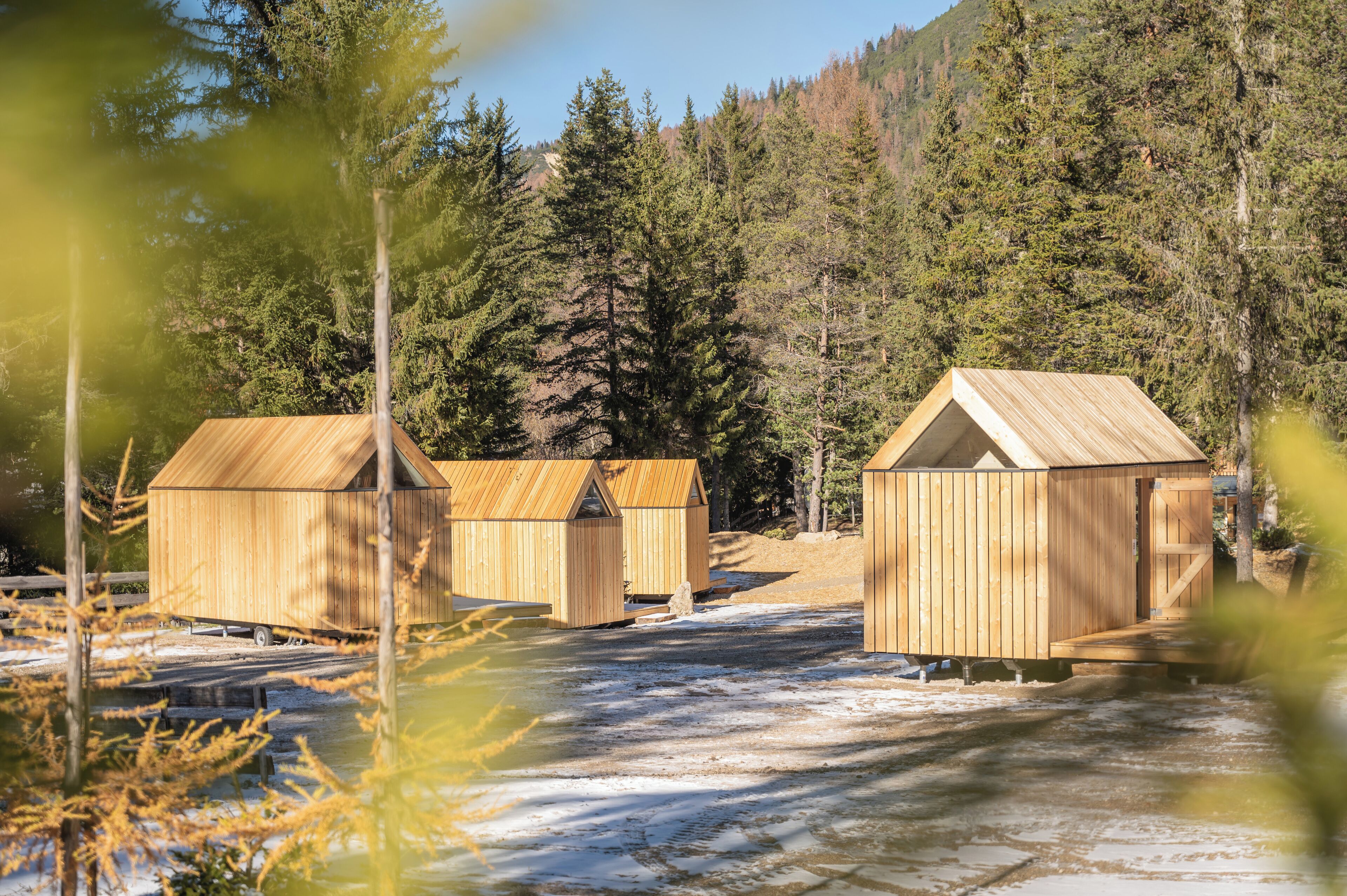 Three wooden cabins with pitched roofs, surrounded by a forested area and a body of water.