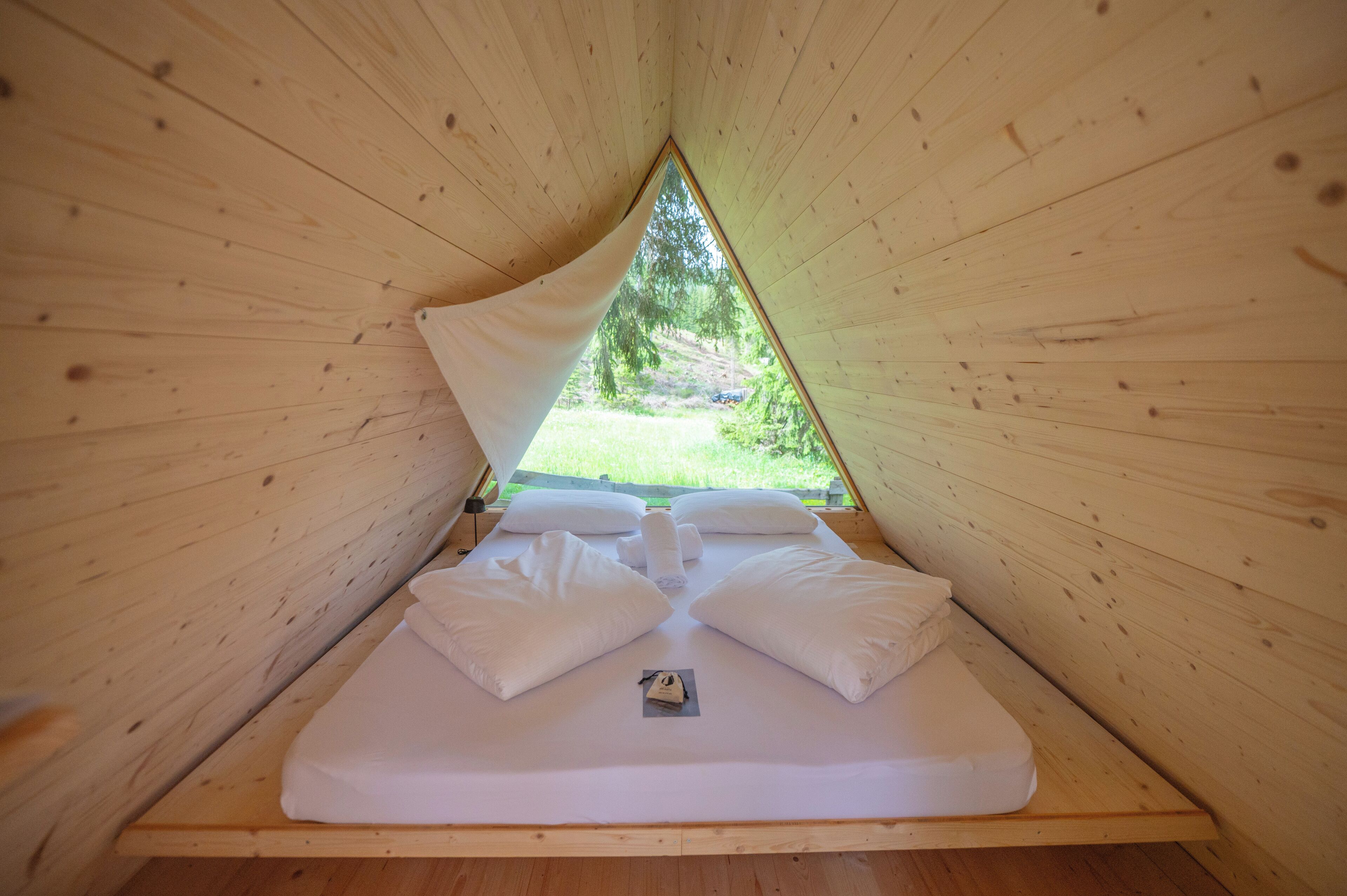 A triangular wooden tent with a bed, pillows, and a blanket.