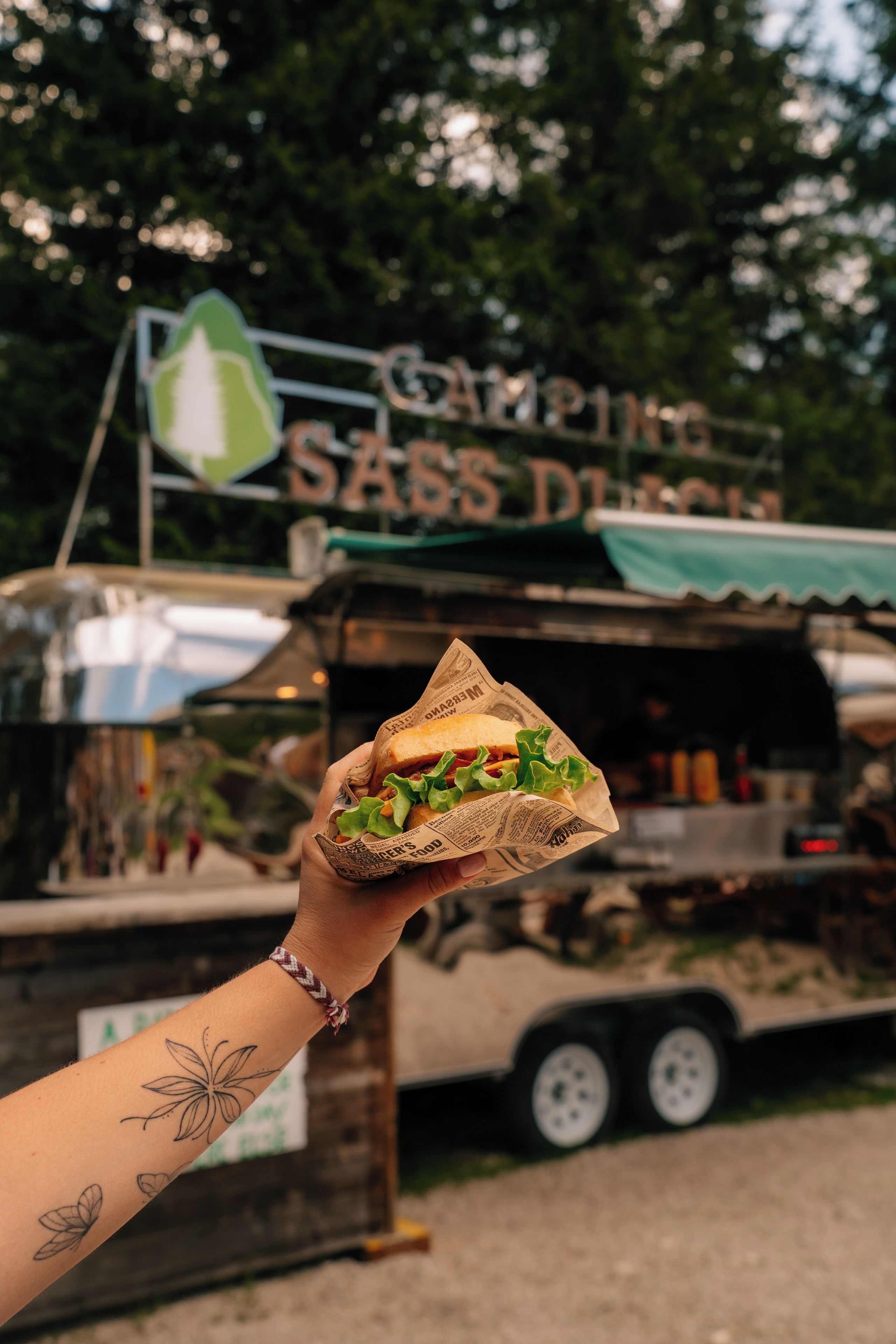 A hand holding a sandwich with a sign in the background that reads 'Camping SASS DINER'.