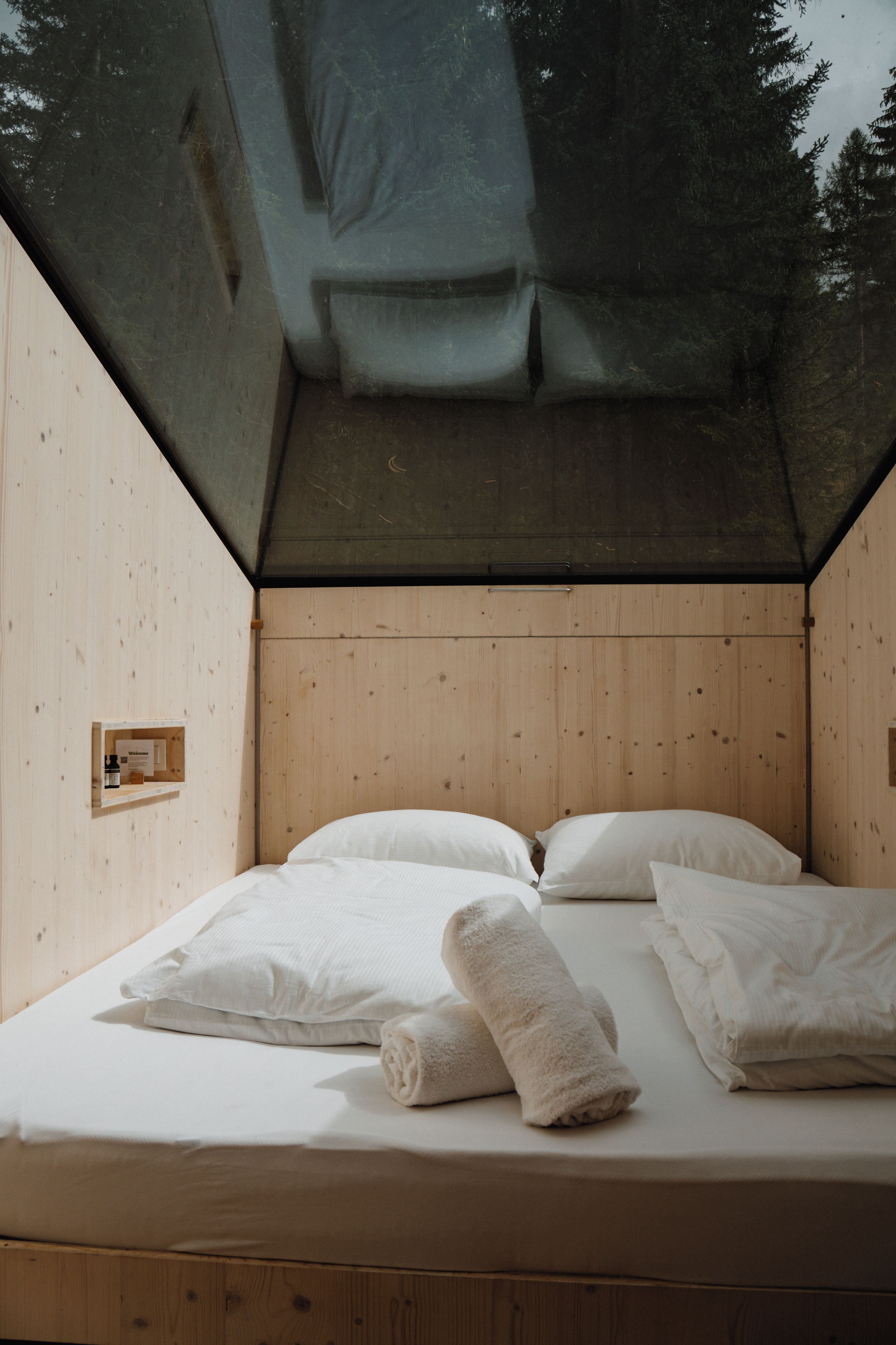 A bed with white linens and pillows, a rolled towel, and a small welcome sign in a wooden cabin with a sloped ceiling.