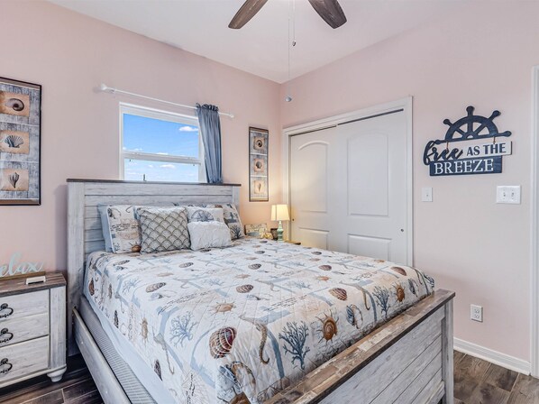 A bedroom with a bed, a nightstand, a window with curtains, a door, and wall decorations including a nautical-themed sign. - Twin Palms Rental 4 Bedroom Home (Rockport)