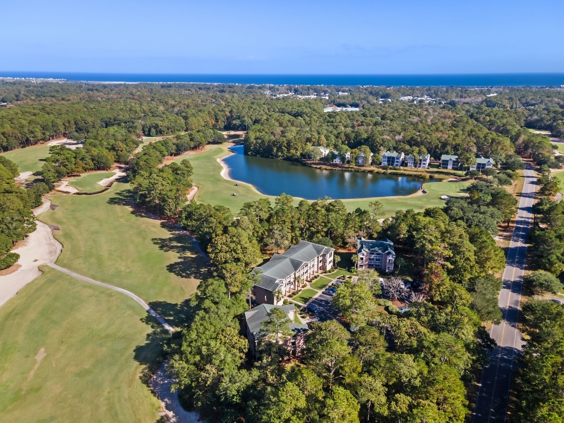 An aerial view of a golf course with a lake, surrounded by dense forests and a row of buildings.