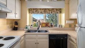 A kitchen with a stove, sink, and dishwasher, a window with a floral curtain, and cabinets. - Hillsider 7 2 Bedroom Townhouse (Steamboat Springs)