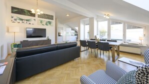 A spacious living area with a large black sofa, a dining table with chairs, and a TV mounted on the wall.
