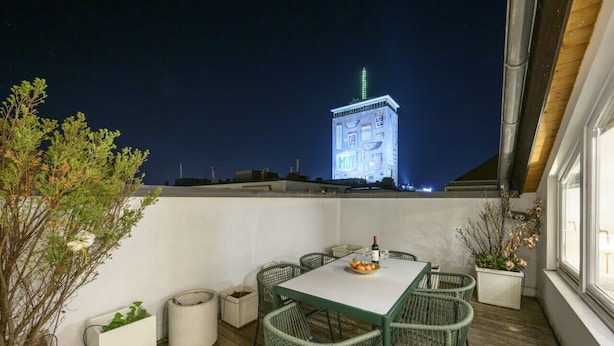 A rooftop terrace with a table and chairs, potted plants, and a view of a lit building in the background.