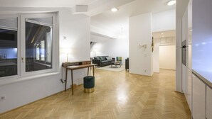 A spacious living area with a wooden floor, a sofa, a coffee table, and a lamp. There is a window with blinds, a door, and a view of the outside.