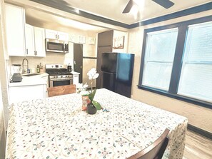 Une cuisine avec des meubles blancs, un four à micro-ondes, une cuisinière, un réfrigérateur et une table à manger recouverte d’une nappe à motifs floraux.