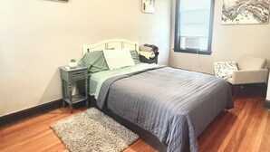 Un dormitorio con cama, mesita de noche, silla, ventana y ventilador de techo.