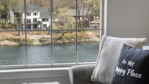 A view through a window of a river with houses in the background, a chair with a pillow, and a window sill.
