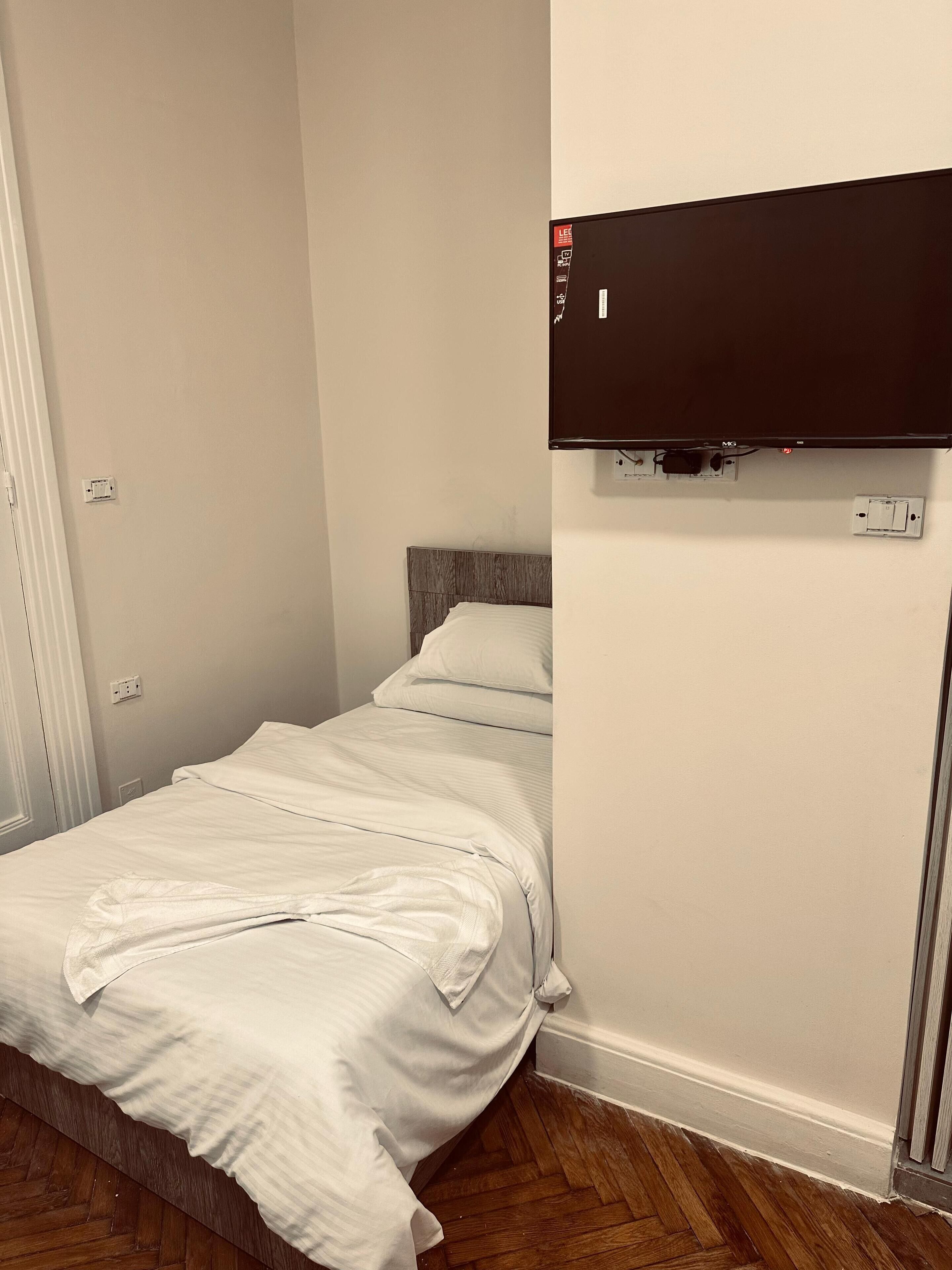 Een klein, keurig opgemaakt bed met witte lakens in een kamer met een houten vloer en een flatscreen televisie aan de muur.