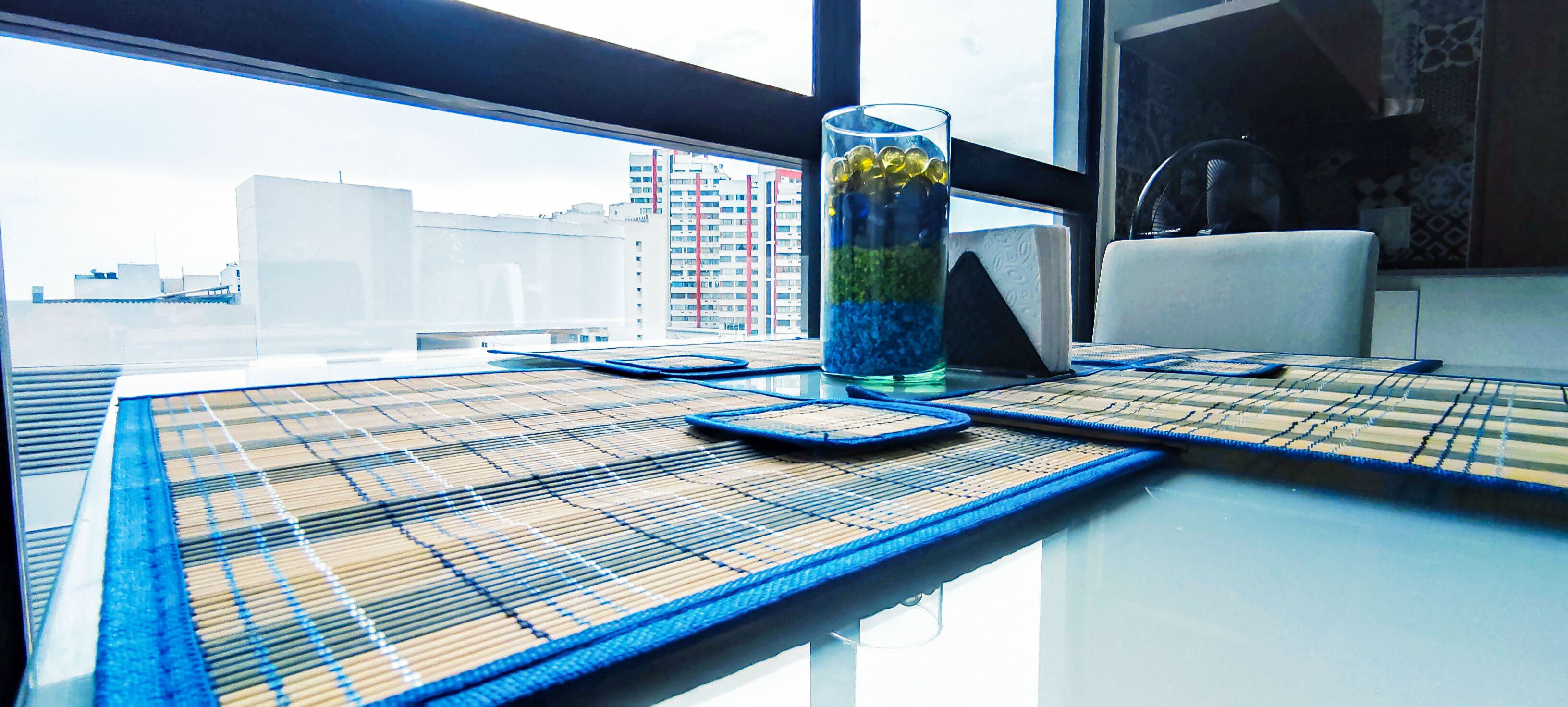 Une table sur laquelle on aperçoit un verre rempli de perles superposées, un téléphone intelligent et un ventilateur, dans une pièce d’où l’on peut voir des bâtiments à travers une fenêtre.