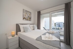 A modern bedroom with a large bed, a bedside lamp, a view of the city through a sliding glass door, and a painting on the wall. -  Smash 2 - Two-bedroom Apartments (Novalja)