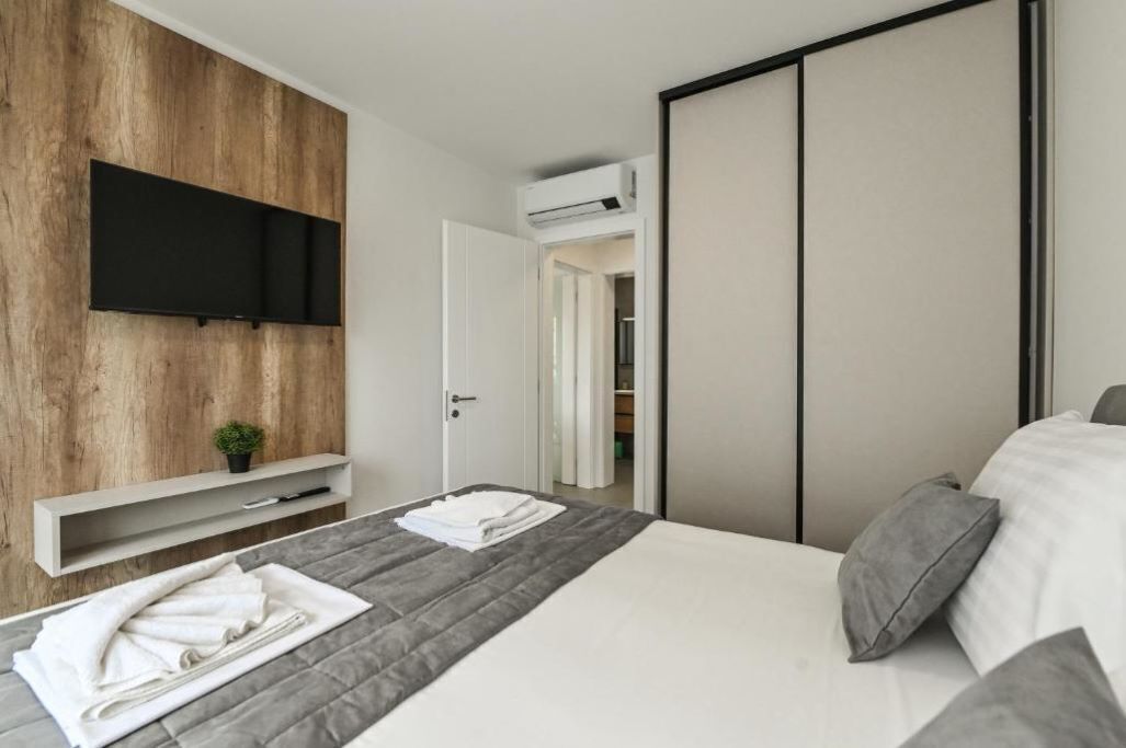 A modern hotel room with a large bed, a flat-screen TV mounted on a wooden wall, a small plant on a shelf, and a wardrobe with sliding doors.