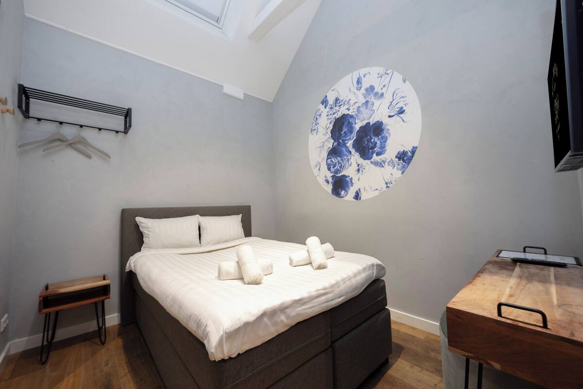 A bedroom with a bed, a wall-mounted heater, a wooden side table, and a circular floral artwork on the wall.