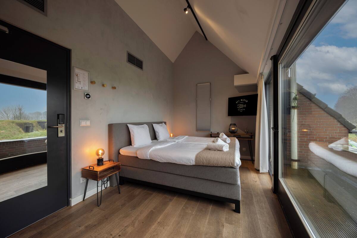 A modern bedroom with a large bed, a bedside table with a lamp, a window with a view of the outside, and a small desk with a chair.