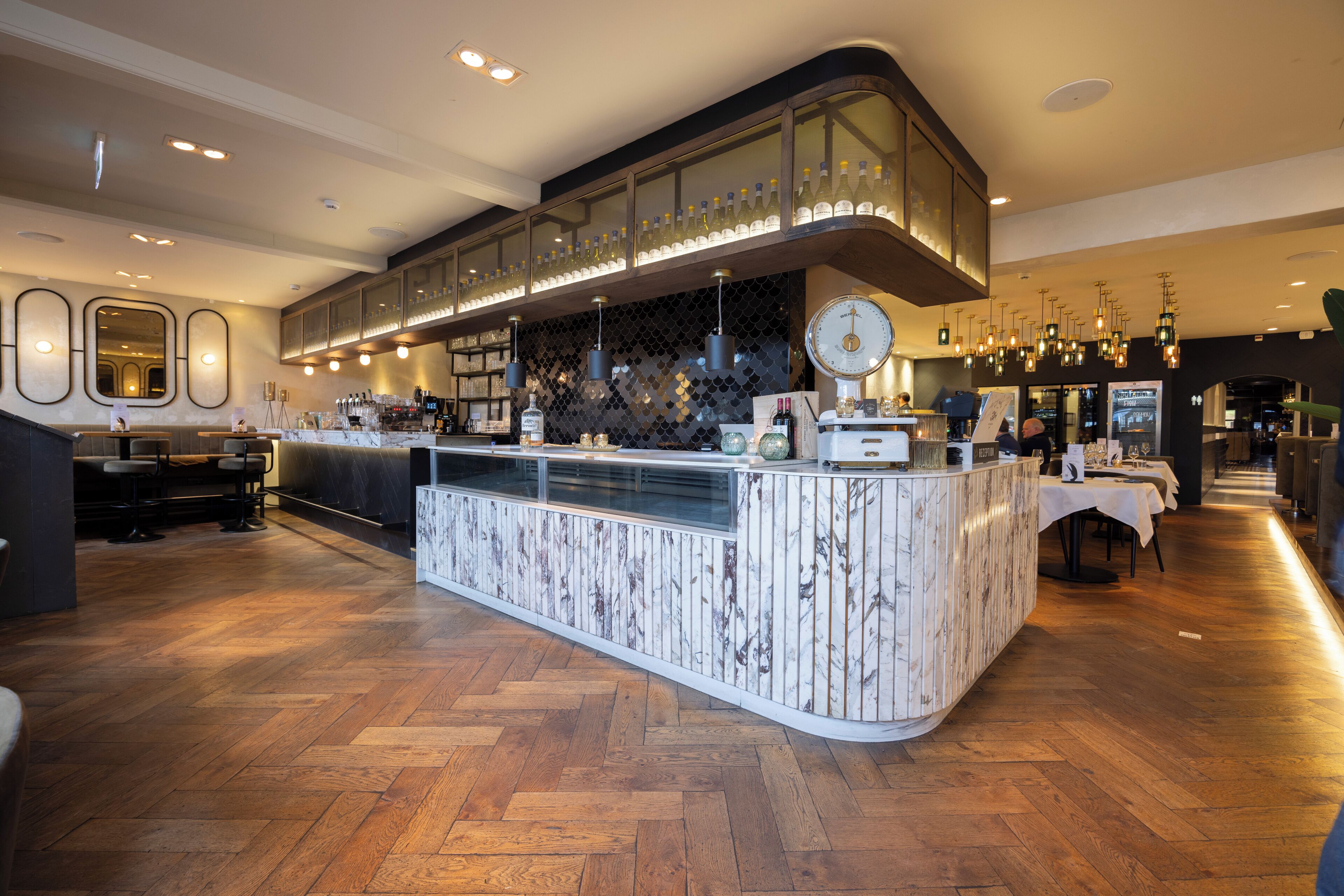 A modern bar area with a marble counter, overhead shelves with bottles, and pendant lighting.