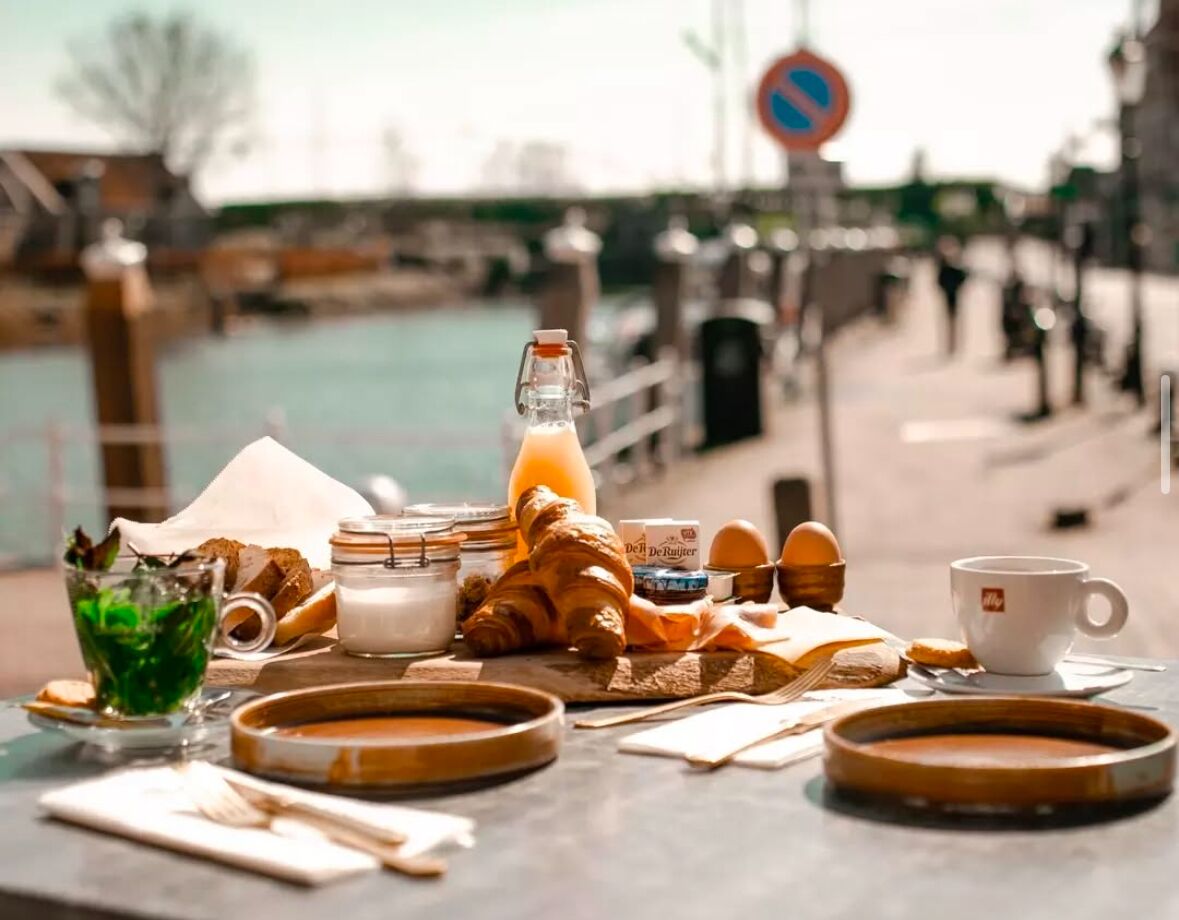 A breakfast setup with croissants, eggs, and a bottle of orange juice on a table by a waterfront.