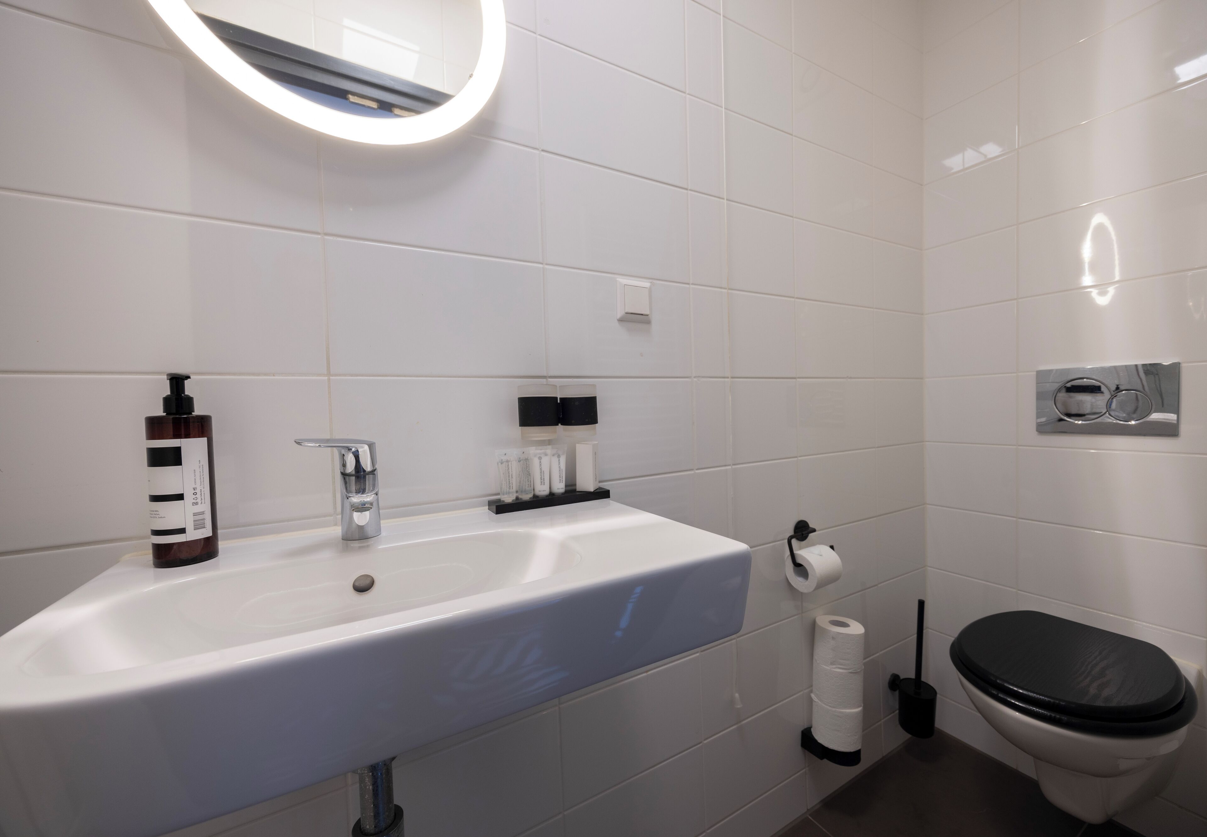 A modern bathroom with a white sink, a mirror with a circular light, a toilet, and a trash bin.