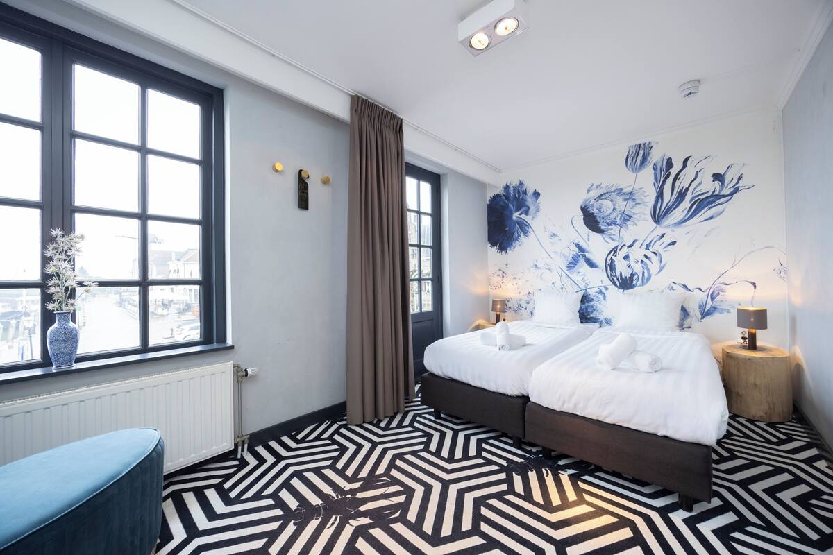 A hotel room with a large window, a bed with white linens, a patterned floor, and a wall mural.