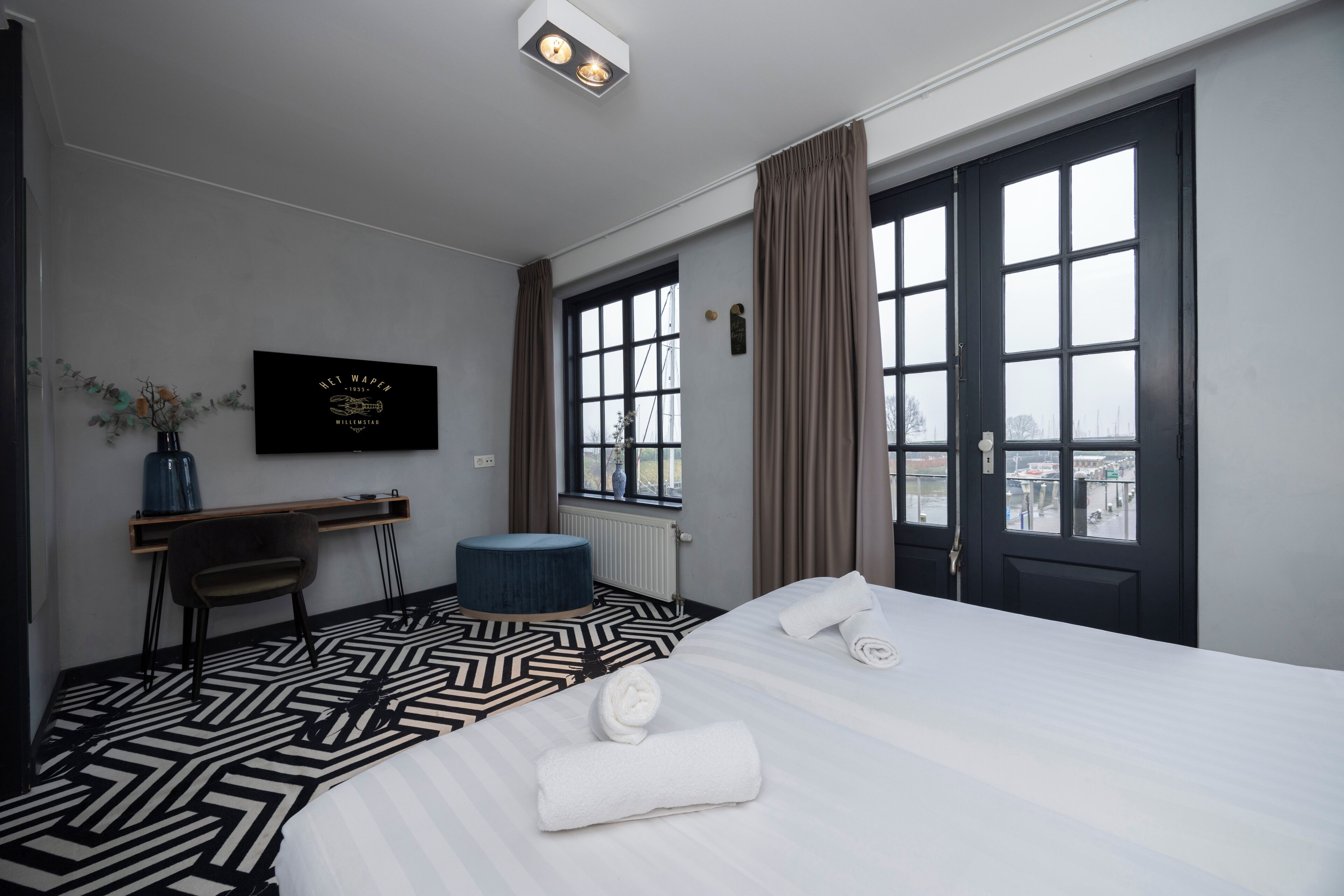 Camera d'albergo con un letto grande, una scrivania, una sedia, un poggiapiedi rotondo, una televisione e una finestra con tende.