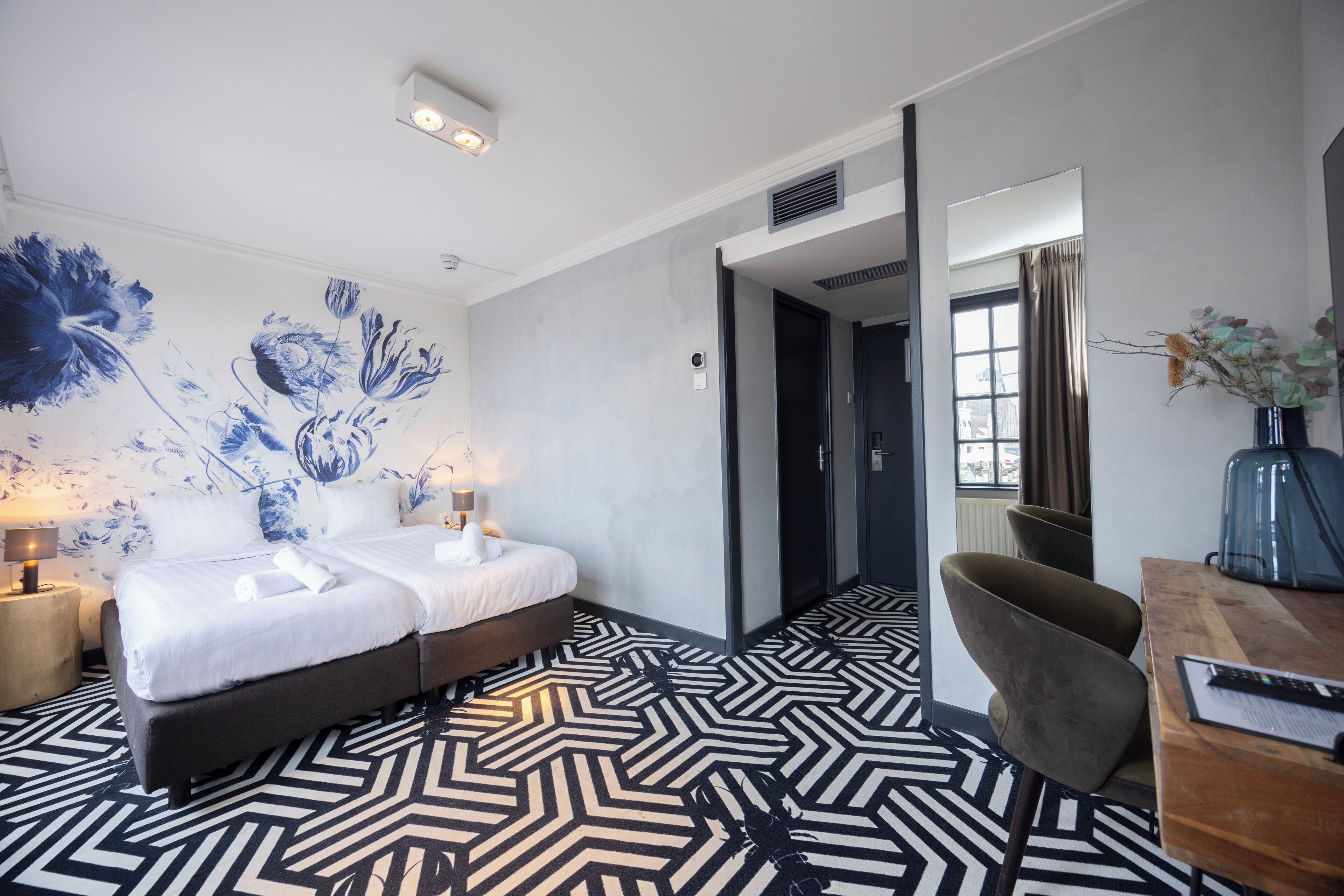 A hotel room with a bed, a desk, a chair, a mirror, and a wall with a floral mural.