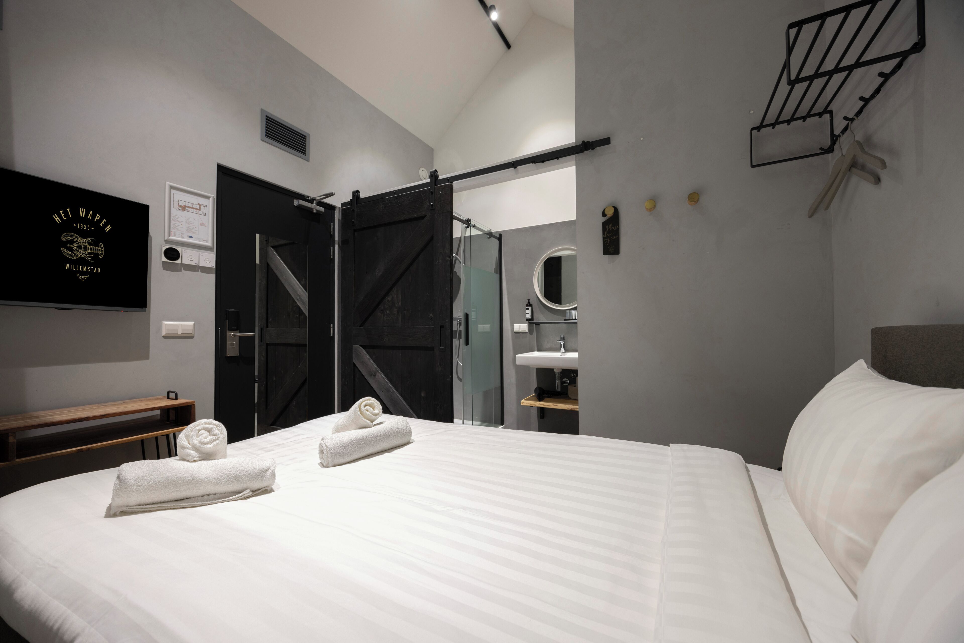 Quarto de hotel moderno com uma cama grande, um banco de madeira, uma porta deslizante preta, um banheiro com pia e espelho, e um porta-toalhas fixado na parede.