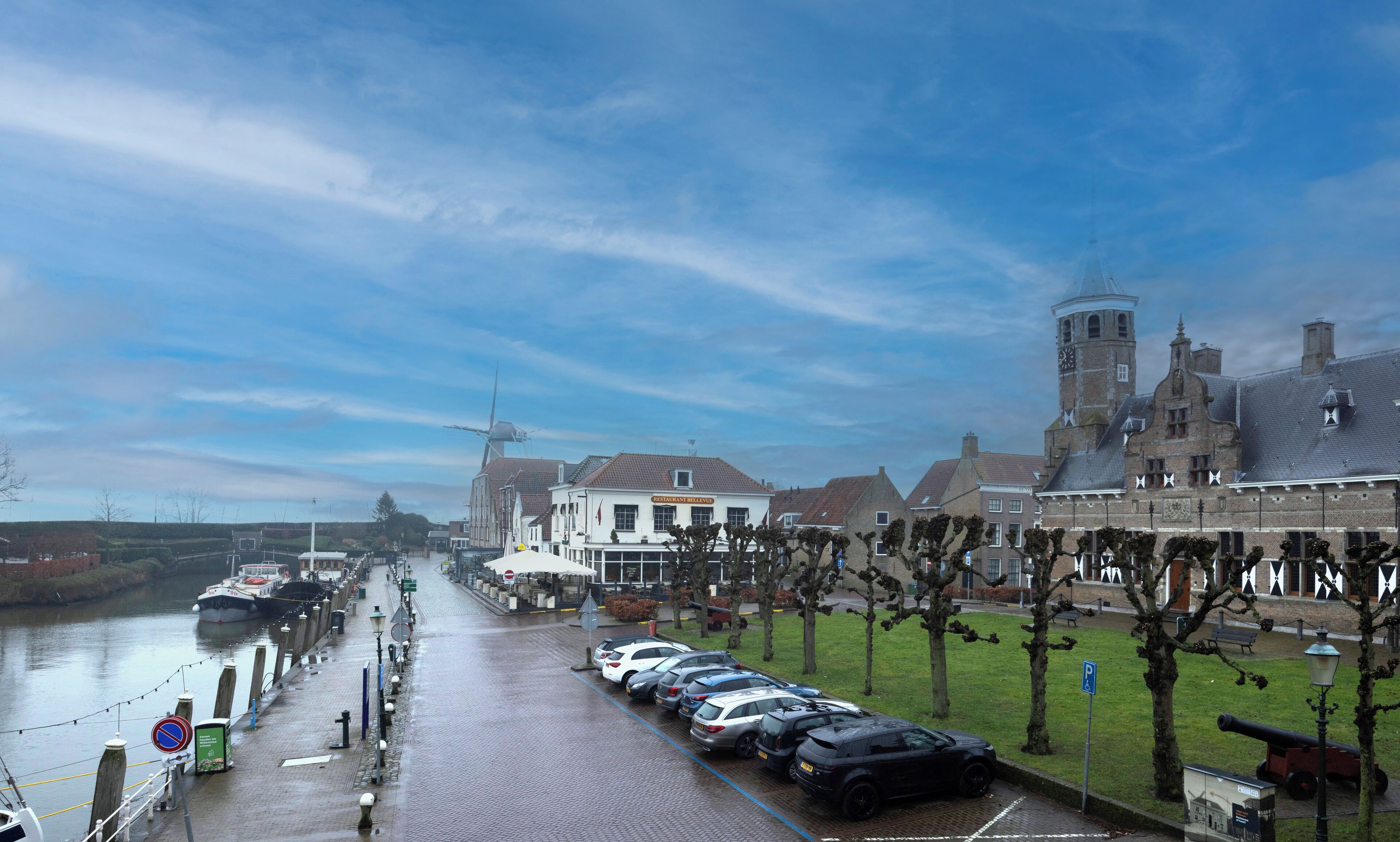 Udsigt over en flod til en historisk by, med parkerede biler, en kanal og traditionelle bygninger.