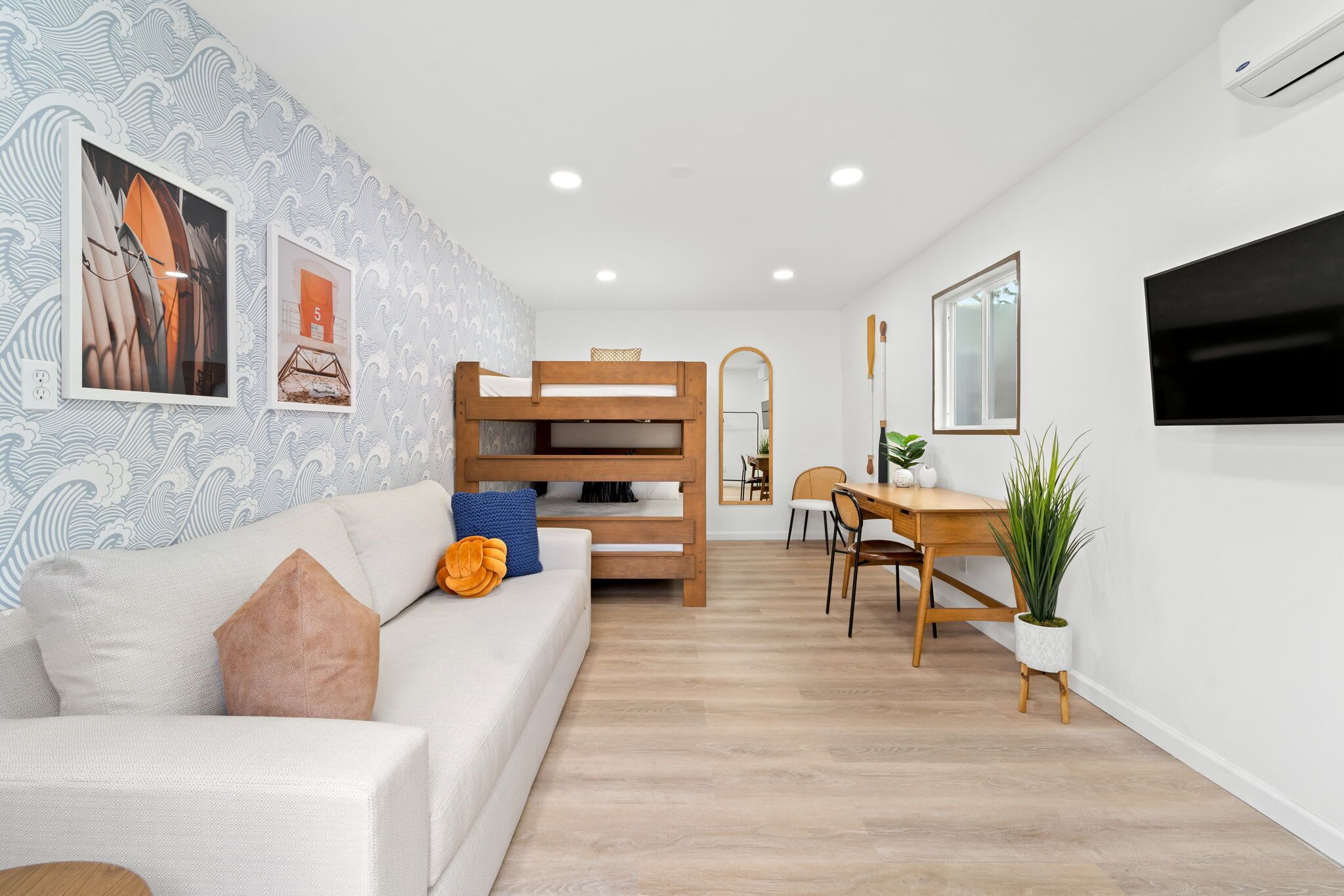 A modern living room with a white sofa, a wooden bunk bed, a dining table with chairs, and a TV mounted on the wall.