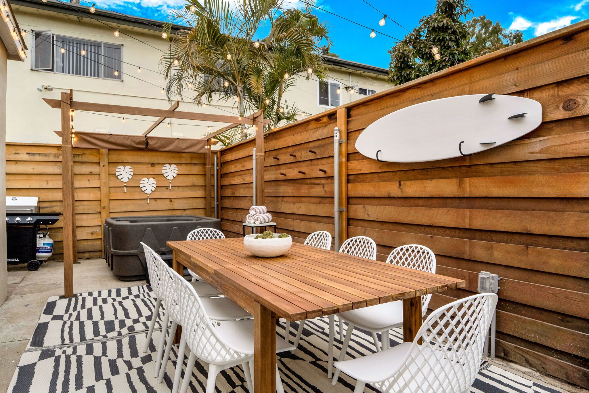 Een houten terras met een eettafel en stoelen, een barbecue en een surfplank aan de muur.