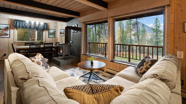 A cozy living room with a sofa, a coffee table, and a fireplace. Large windows offer a view of the forest.