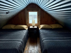 A small attic bedroom with two beds, a nightstand, and a window. - Raven Cove - Tiny House with a Twist (Findley Lake)