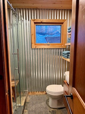 A compact bathroom with a toilet, a small window, and a shower area. - Raven Cove - Tiny House with a Twist (Findley Lake)