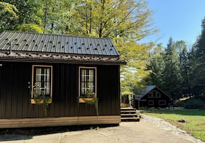 A rustic cabin with a metal roof, two windows with wooden frames, and a small porch area with a planter. - Raven Cove - Tiny House with a Twist (Findley Lake)