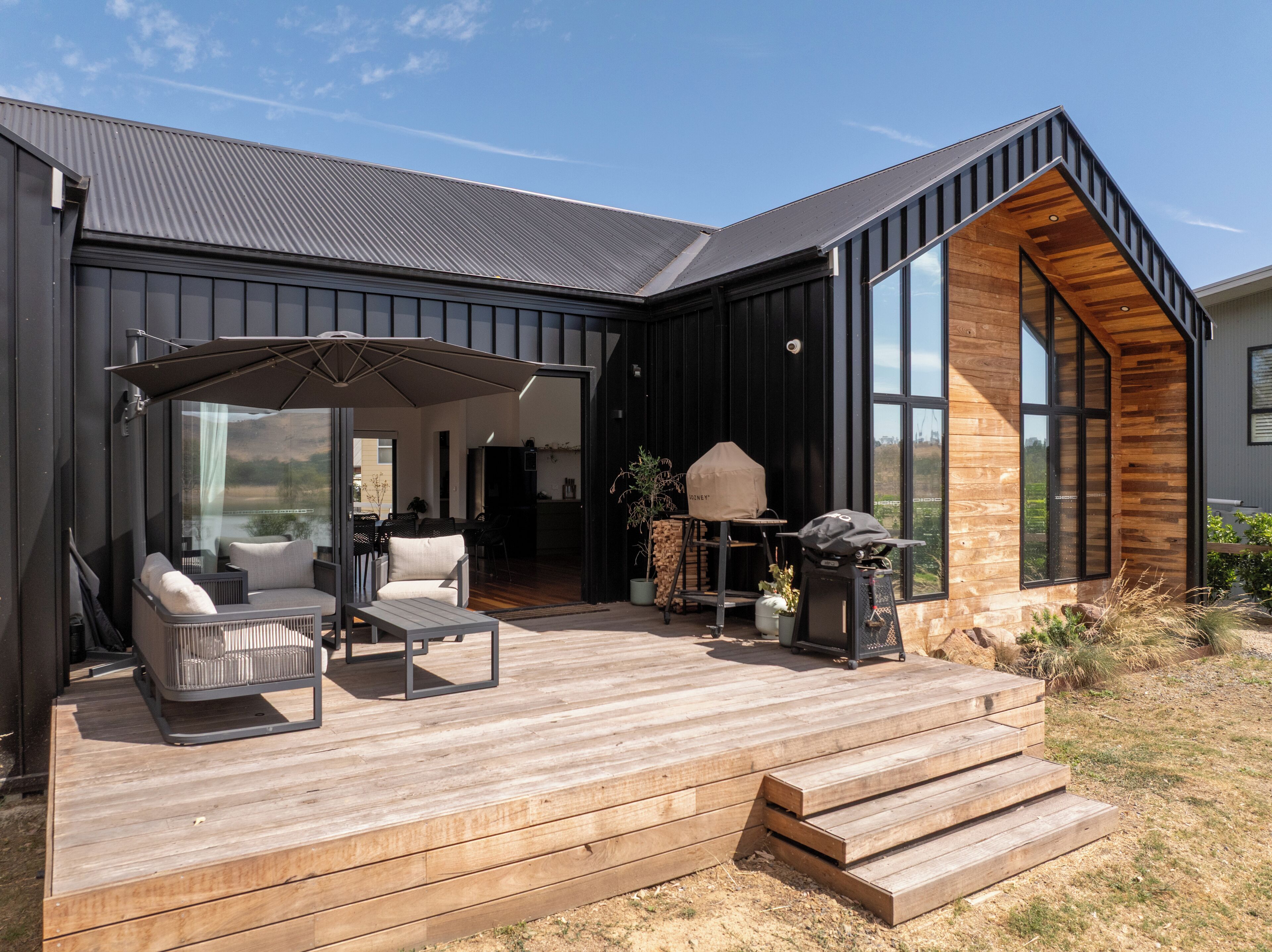 Ein modernes Haus mit einer Holzterrasse, Sitzgelegenheiten im Freien und einem großen Fenster mit Blick auf die Landschaft.